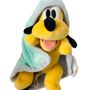 Puede incluir: Un juguete de peluche de Pluto el perro con una manta con capucha azul y blanca. El juguete está sentado con la lengua fuera.