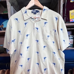 May include: A light tan Polo shirt with a blue marlin pattern. The shirt has short sleeves, a collared neckline, and a single chest pocket. The Polo by Ralph Lauren label is visible.