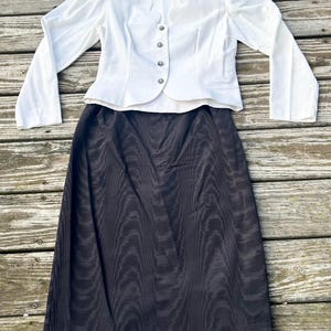 May include: A two-piece outfit featuring a white long-sleeve jacket with decorative buttons and a black skirt. The jacket has a scalloped neckline and the skirt is knee-length. The outfit is displayed on a wooden surface.