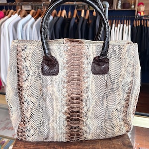 May include: A large, rectangular handbag with a snakeskin pattern in shades of beige, brown, and cream. The bag has dark brown leather handles and accents. The bag is sitting on a wooden surface.