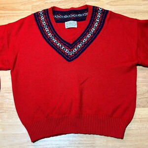 May include: A red V-neck sweater with long sleeves. The neckline and cuffs feature a navy blue, white, and red geometric pattern. The sweater has a ribbed hem and cuffs. A small tag is visible near the neckline.