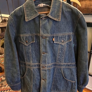 May include: A vintage denim jacket with a button-up front, two chest pockets, and two lower pockets. The jacket has a classic collar and is a medium blue wash. The jacket is hanging on a wooden hanger.