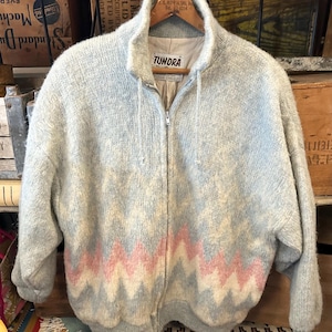 May include: A light gray zip-up jacket with a pink and white zigzag pattern across the lower portion. The jacket has a high collar and the brand name "TUNDRA" is visible on the label. The jacket is hanging on a wooden hanger.