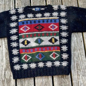 May include: A black sweater with a colorful geometric pattern. The sweater features a central design with red, blue, green, and yellow accents, framed by a border of white snowflake-like shapes. The sleeves have similar patterns.