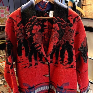 May include: A red wool jacket with a black and white design featuring figures and geometric patterns. The jacket has a notched collar, two buttons, and a blue and black trim. The jacket is hanging on a wooden hanger.