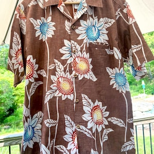 May include: Brown short-sleeve button-down shirt with a sunflower pattern. The sunflowers have white petals, red and yellow centers, and blue accents. The shirt has a chest pocket and is hanging on a wooden hanger.