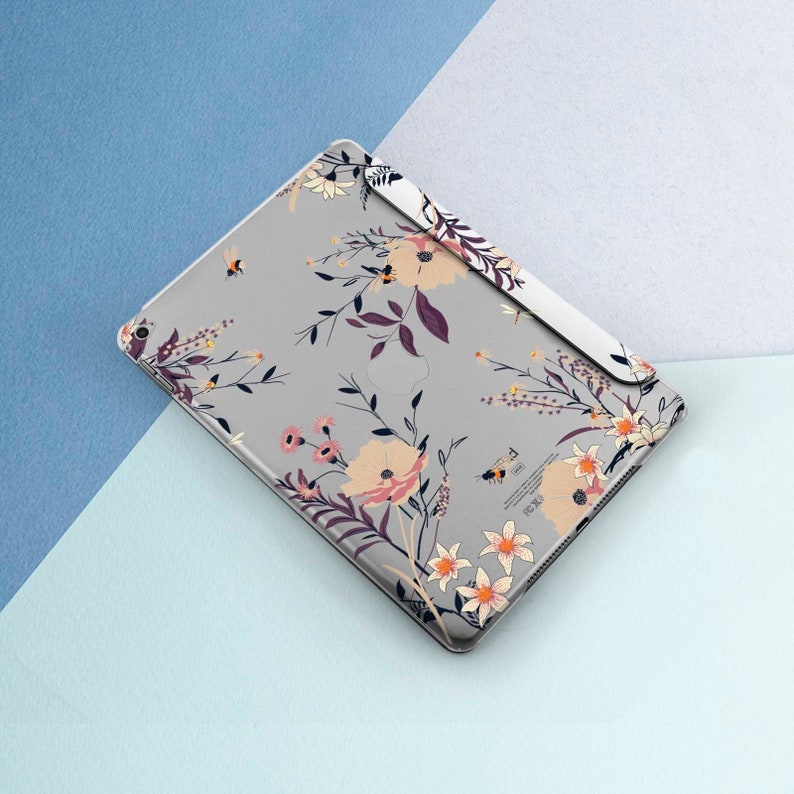 May include: A gray iPad case with a floral pattern featuring pink, yellow, and white flowers, green leaves, and small bees. The case has a cutout for the Apple logo.