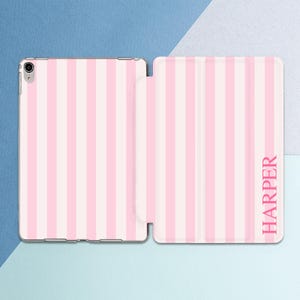 May include: A tablet case featuring a vertical striped pattern in alternating shades of light pink and white. The right side of the folding cover includes the name HARPER printed in a vertical orientation using a bold, dark pink sans-serif font.