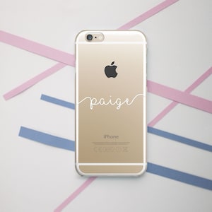 May include: A gold iPhone case with a white inscription that says "paige" in cursive. The case is clear and shows the Apple logo on the back.