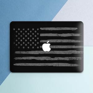 May include: Black laptop skin featuring a distressed American flag design with a white Apple logo. The flag has gray stripes and stars, creating a patriotic and modern aesthetic. The skin is rectangular and fits a laptop.