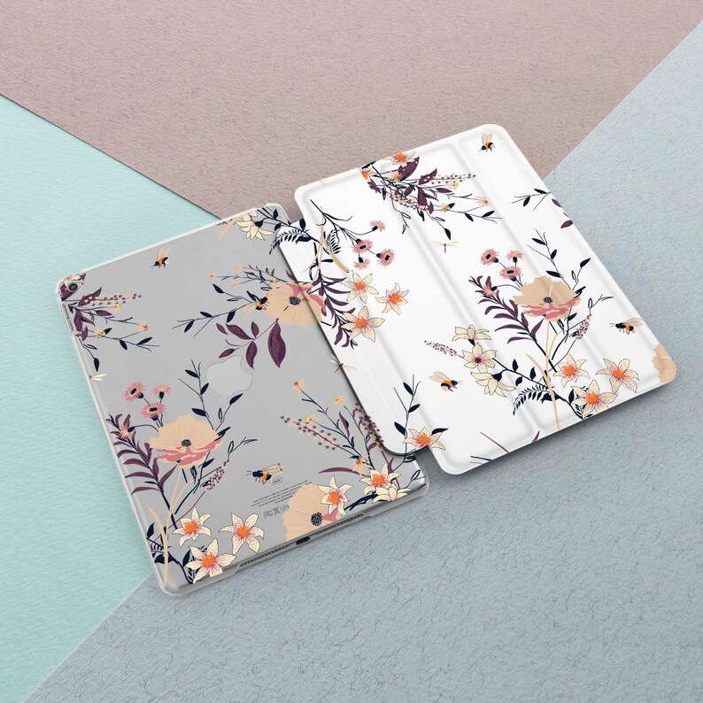 May include: Two floral patterned iPad cases. One case is white with a light pink and yellow floral pattern. The other case is gray with a darker pink and yellow floral pattern. Both cases have a bee design.