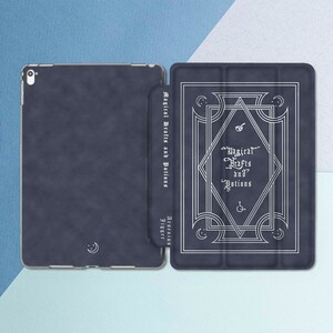 Peut inclure: Une coque pour iPad bleue avec un design décoratif comprenant un cadre argenté et le texte "Magical Crafts and Potions" en argent. La coque est fermée et le dos de la coque présente un motif de croissant de lune.