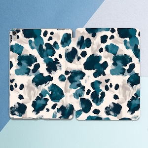 Puede incluir: Funda para tableta con fondo crema y un patrón de formas abstractas en azul verdoso y azul marino. La funda está abierta, revelando el interior y el dispositivo que protege.