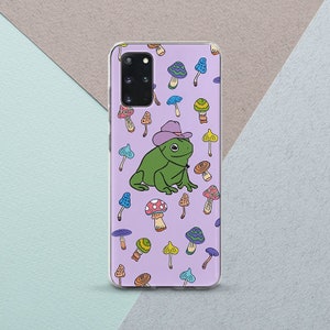以下が含まれることがあります： 紫色の携帯電話ケースに、紫色のカウボーイハットをかぶった緑色のカエルが、カラフルなキノコに囲まれているデザインです。