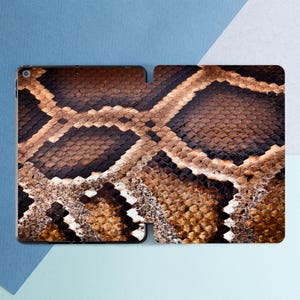 May include: A tablet case featuring a detailed snakeskin pattern in shades of brown, tan, and black. The design mimics the texture and coloration of reptile scales, creating a realistic and naturalistic appearance. The case is open, revealing the interior.