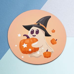 Puede incluir: Un posavasos redondo con un fantasma de dibujos animados con un sombrero de bruja y una calabaza naranja con detalles de estrellas blancas. Hay calabazas y estrellas adicionales dispersas sobre un fondo melocotón.