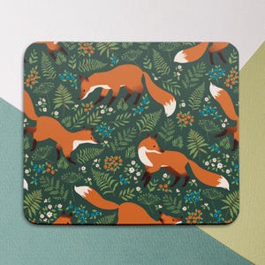 May include: A rectangular mousepad featuring a repeating pattern of orange foxes with white-tipped tails, set against a dark green background filled with ferns, berries, and small white flowers. The design has a whimsical, nature-inspired theme.