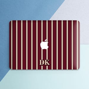 May include: A burgundy and cream striped laptop skin with a white Apple logo and the initials "DK" in a serif font. The skin has a glossy finish and is designed to fit a laptop. The background is a mix of blue and white.