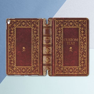 Puede incluir: Una funda para teléfono de cuero rojo con un diseño dorado ornamentado. La funda está dividida en dos secciones, con el lado izquierdo que presenta una columna vertebral dorada con la palabra "TOME" y el lado derecho que presenta el texto "CUSTOM TEXT".