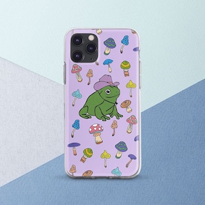May include: A clear phone case with a purple background and a green frog wearing a purple cowboy hat. The frog is surrounded by colorful mushrooms.