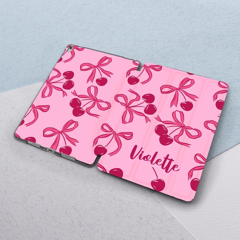 May include: Pink tablet case with a cherry and bow pattern. The case has the name "Violette" printed on it.
