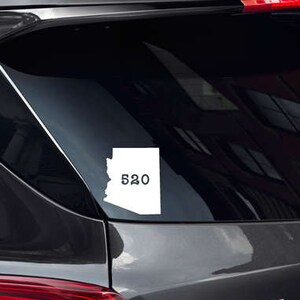 Op de afbeelding: Een witte vinyl sticker van de staat Arizona met het nummer 620 binnen de omtrek van de staat. De sticker zit op de achterruit van een zwarte auto.