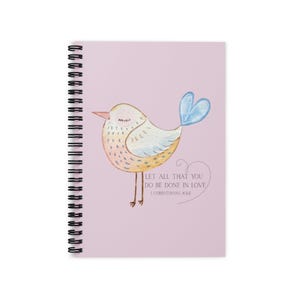 May include: A spiral-bound notebook with a light pink cover featuring a watercolor illustration of a bird. The bird is yellow, white, and blue, with the text "LET ALL THAT YOU DO BE DONE IN LOVE" and "1 CORINTHIANS 16:14".