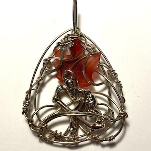 May include: A silver-toned wire-wrapped pendant featuring a central metallic hockey player figure in a skating pose. Behind the figure is a translucent, reddish-orange maple leaf shape. The entire piece is encased in a triangular wire frame.