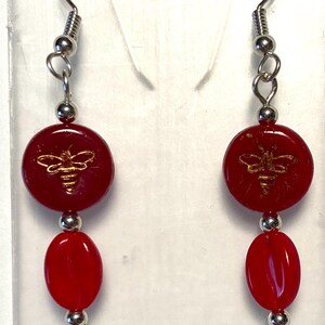 May include: A pair of dangle earrings featuring a red, circular bead with a gold bee design, connected to a red oval bead and silver-colored hardware. The earrings have a hook closure.