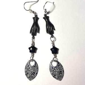 May include: A pair of dangle earrings featuring silver-toned hooks, black hand-shaped charms, small black star-shaped beads, and teardrop-shaped pendants with a white and black circuit board pattern printed on the surface of each piece.