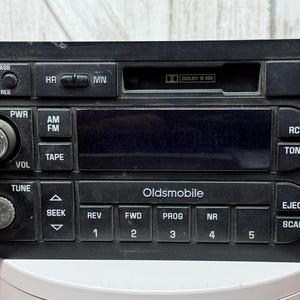 May include: A black Oldsmobile car radio head unit featuring a cassette player slot with Dolby NR, a digital display, volume and tuning knobs, and various control buttons labeled AM, FM, TAPE, RCL, TONE, SEEK, REV, FWD, PROG, NR, EJECT, and SCAN.