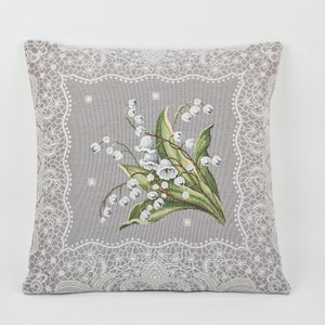 Può includere: Un cuscino grigio con un bordo in pizzo bianco e un motivo floreale di mughetti bianchi su sfondo grigio.