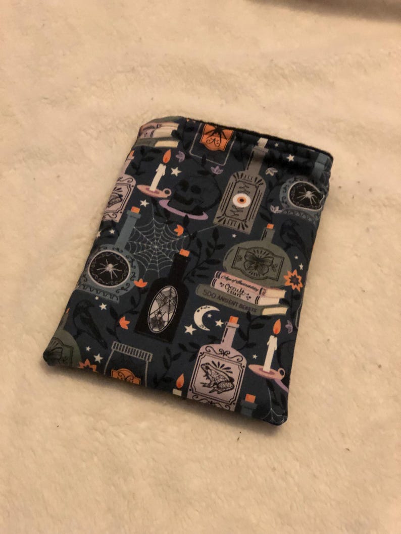 Może przedstawiać: Niebieska, materiałowa torebka z powtarzającym się wzorem butelek z eliksirami, świec i innych czarodziejskich przedmiot&oacute;w. Wz&oacute;r jest w odcieniach niebieskiego, fioletowego, pomarańczowego i białego.