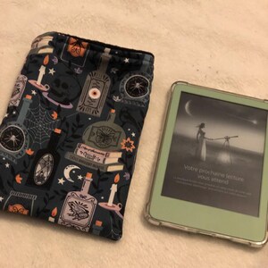 Może przedstawiać: Czarno-niebieska osłonka na czytnik e-book&oacute;w z przerażającym wzorem na Halloween, przedstawiającym butelki, świece, pająki i czaszkę. Osłonka leży obok białego czytnika e-book&oacute;w w zielonym etui. Ekran czytnika e-book&oacute;w wyświetla tekst "Votre prochaine lecture vous attend".