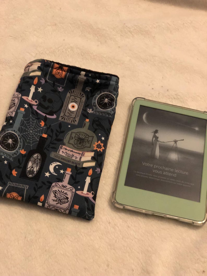 Może przedstawiać: Czarno-niebieska osłonka na czytnik e-book&oacute;w z przerażającym wzorem na Halloween, przedstawiającym butelki, świece, pająki i czaszkę. Osłonka leży obok białego czytnika e-book&oacute;w w zielonym etui. Ekran czytnika e-book&oacute;w wyświetla tekst "Votre prochaine lecture vous attend".