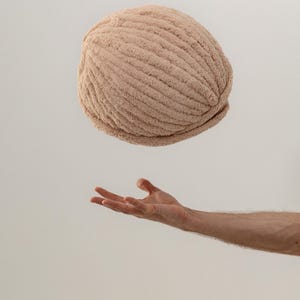 Op de afbeelding: Een ronde, beige, getextureerde muts met een dik geribbeld breipatroon. De muts heeft een zacht, pluizig uiterlijk met een consistente lichtbruine kleur over het hele oppervlak, met een eenvoudige ronde vorm en een omgeslagen rand.