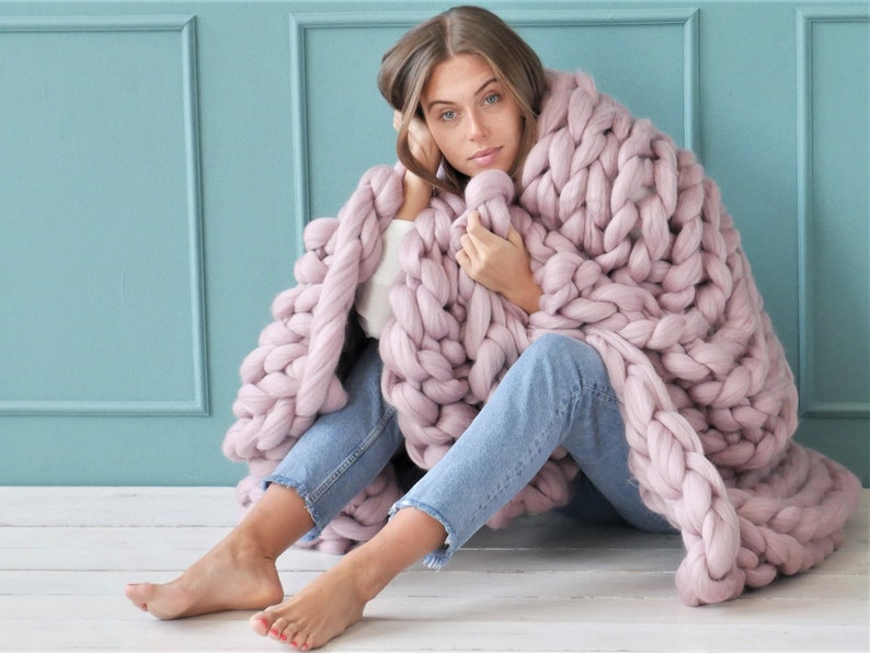 May include: A chunky knit blanket in a soft mauve color. The blanket is made of thick, oversized yarn, creating a cozy and textured appearance. The blanket is draped over a person wearing blue jeans and a white top, sitting against a light blue wall.
