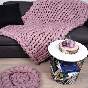 May include: A pink chunky knit throw blanket draped over a gray couch with a black throw pillow. The blanket is also used as a decorative pillow on the floor. A small white side table with a wire base holds a few decorative items.