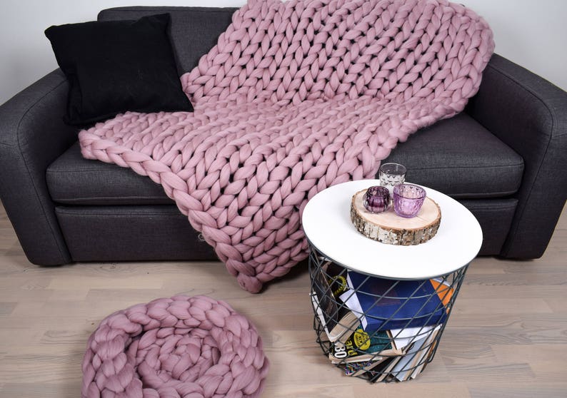 May include: A pink chunky knit throw blanket draped over a gray couch with a black throw pillow. The blanket is also used as a decorative pillow on the floor. A small white side table with a wire base holds a few decorative items.