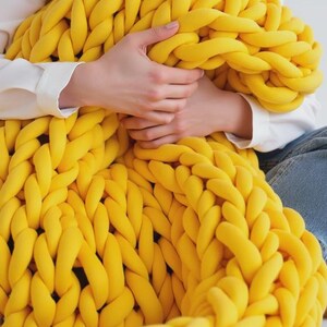 Op de afbeelding: Een grote, dikgebreide deken in een levendige gele kleur. De deken is gemaakt van dik, zacht garen en heeft een getextureerde, gevlochten uitstraling. De deken wordt vastgehouden door een persoon die een wit overhemd en een blauwe jeans draagt. Een gezellig woondecoratie-item.
