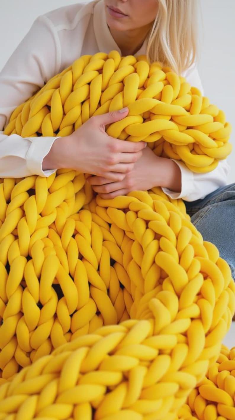 Op de afbeelding: Een grote, dikgebreide deken in een levendige gele kleur. De deken is gemaakt van dik, zacht garen en heeft een getextureerde, gevlochten uitstraling. De deken wordt vastgehouden door een persoon die een wit overhemd en een blauwe jeans draagt. Een gezellig woondecoratie-item.