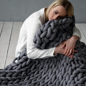 May include: A large, chunky-knit grey blanket. The blanket is made of thick yarn and has a soft, textured appearance. The blanket is draped over a person, suggesting warmth and comfort. The blanket is a home decor item.