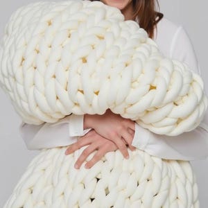 Op de afbeelding: Een grote, ivoorkleurige, dik gebreide deken. De deken is gemaakt van dik, zacht garen en heeft een textuur. De oversized plaid wordt vastgehouden door een persoon die een wit overhemd draagt.