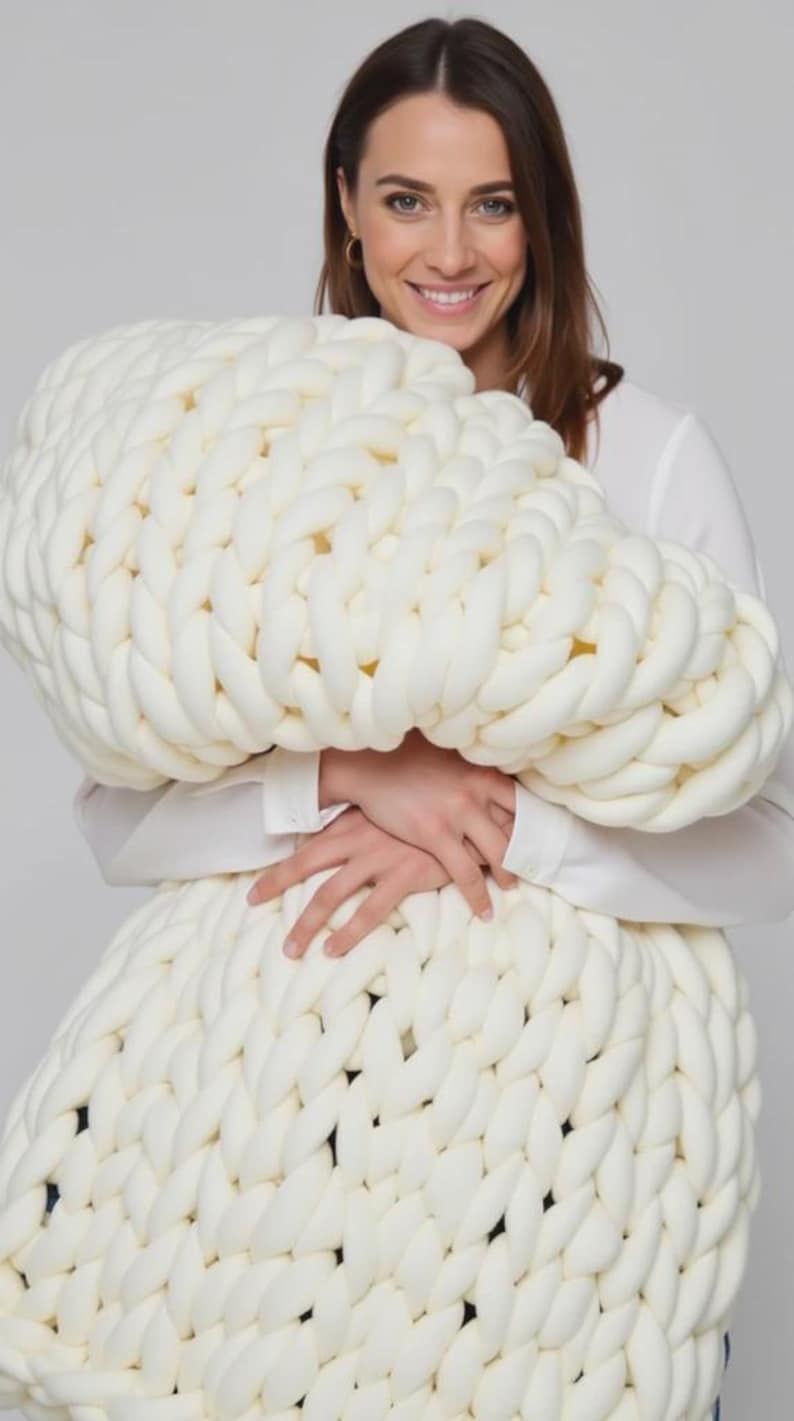 Op de afbeelding: Een grote, ivoorkleurige, dik gebreide deken. De deken is gemaakt van dik, zacht garen en heeft een textuur. De oversized plaid wordt vastgehouden door een persoon die een wit overhemd draagt.