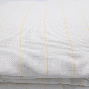 May include: Folded white fabric with vertical yellow stripes. The fabric has a textured appearance, with the stripes evenly spaced across the surface. The material appears to be soft and possibly linen or cotton.
