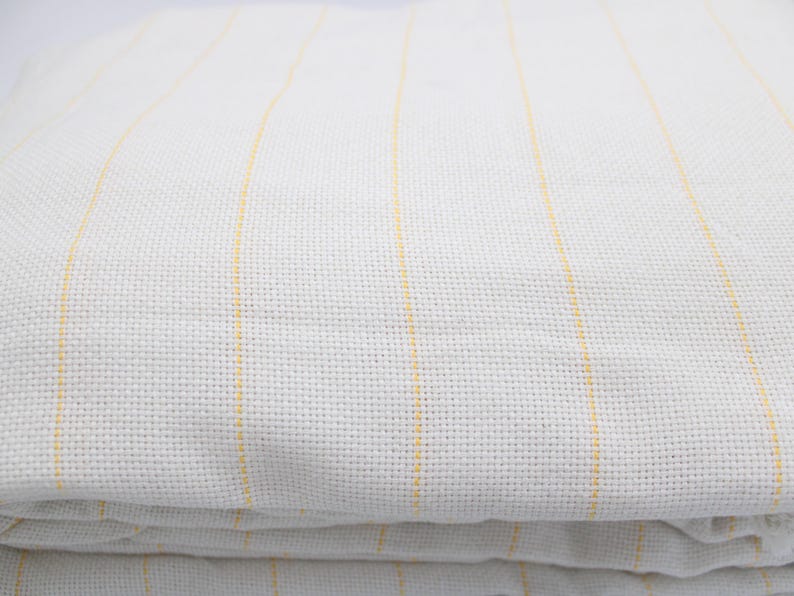 May include: Folded white fabric with vertical yellow stripes. The fabric has a textured appearance, with the stripes evenly spaced across the surface. The material appears to be soft and possibly linen or cotton.