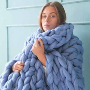 May include: A large, chunky-knit blanket in a soft periwinkle blue. The blanket is made of thick yarn, creating a textured, cozy appearance. The oversized knit design is wrapped around the shoulders, suggesting warmth and comfort. The background is a light blue wall.