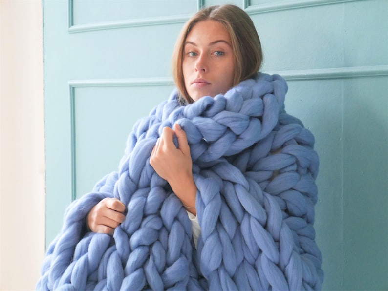 May include: A large, chunky-knit blanket in a soft periwinkle blue. The blanket is made of thick yarn, creating a textured, cozy appearance. The oversized knit design is wrapped around the shoulders, suggesting warmth and comfort. The background is a light blue wall.