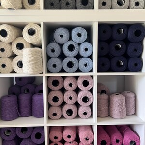May include: A white, multi-compartment shelf filled with yarn in various colors. The yarn is arranged in neat rows, with shades of white, gray, blue, purple, and pink. The yarn is wound into cones and balls, perfect for knitting and crochet projects.