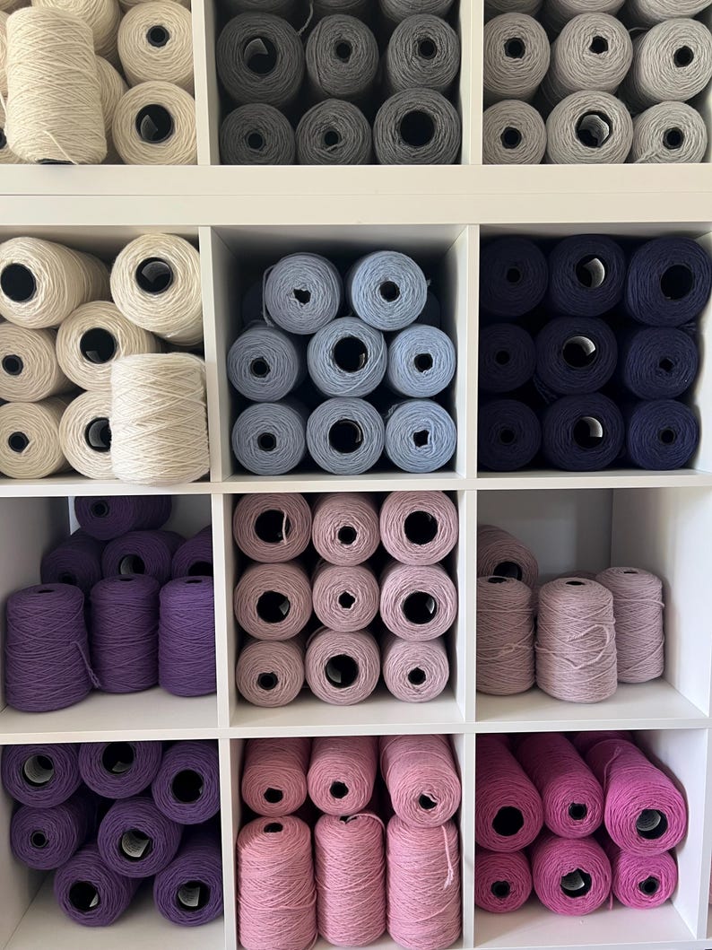 May include: A white, multi-compartment shelf filled with yarn in various colors. The yarn is arranged in neat rows, with shades of white, gray, blue, purple, and pink. The yarn is wound into cones and balls, perfect for knitting and crochet projects.